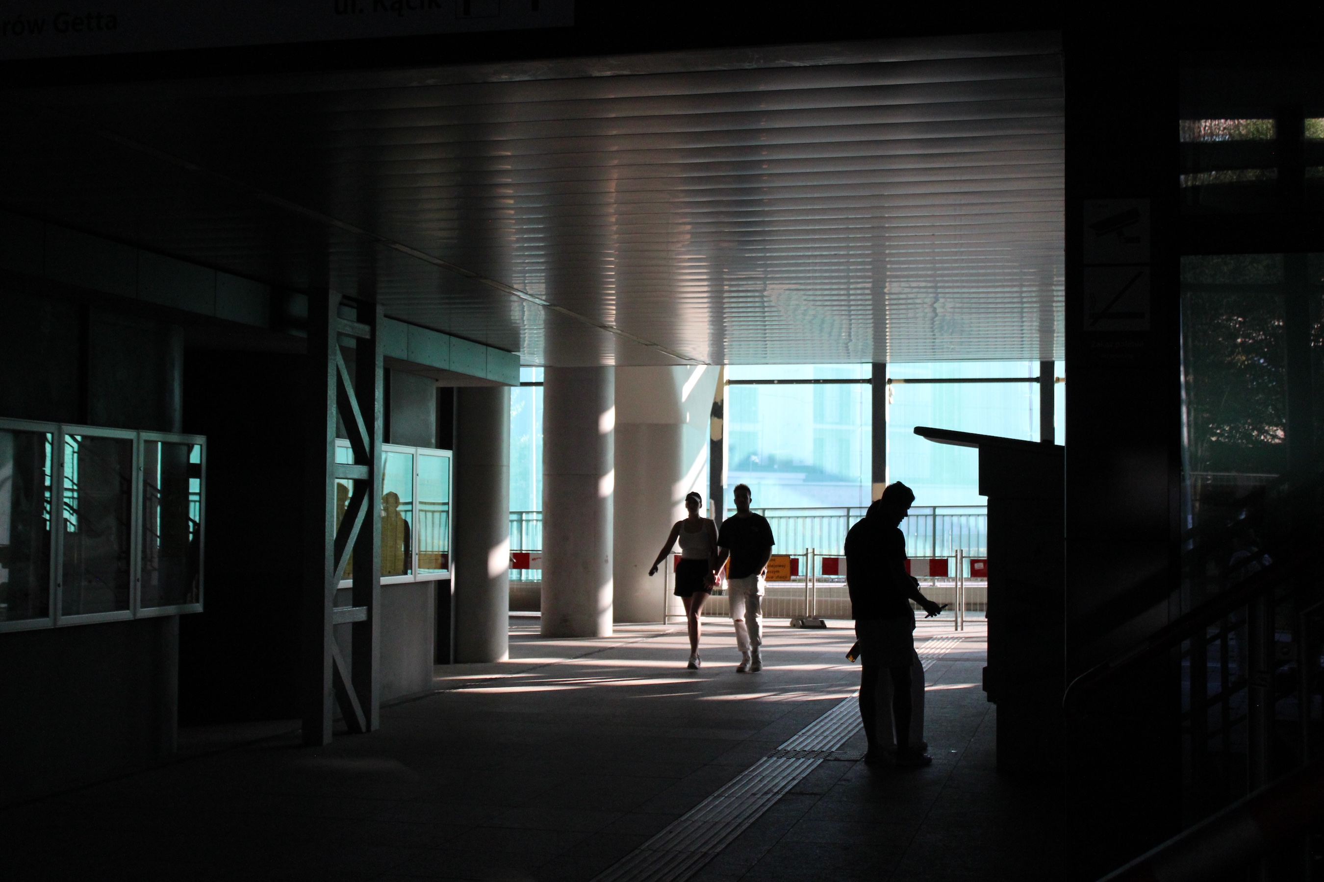 An underpass at the train station. Huge glass windows on the opposite end cast light on some silhouettes of people walking through and other buying tickets. It's otherwise unlit.